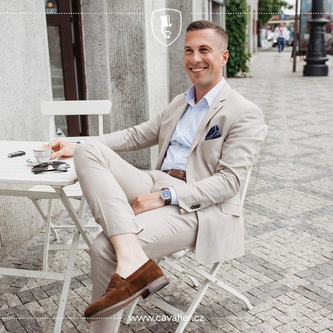 Usměvavý muž v béžovém lněném obleku na míru Cavalier sedící v letní kavárně jako příklad uvolněné elegance a prodyšného materiálu