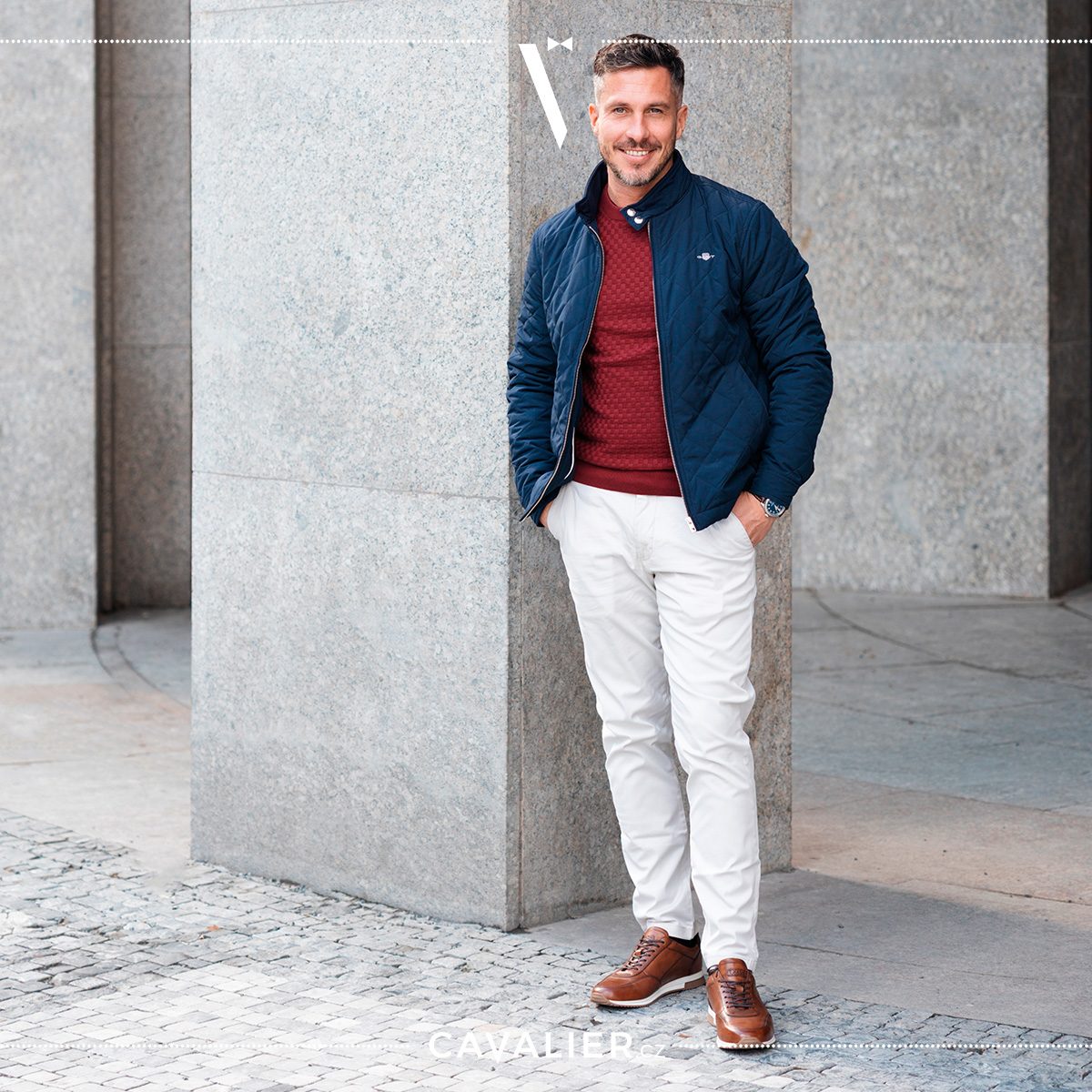 Usměvavý muž v ležérním smart casual outfitu – tmavě modrá prošívaná bunda, vínový pletený svetr a světlé kalhoty na míru Cavalier ukazují stylové vrstvení pro chladnější dny.