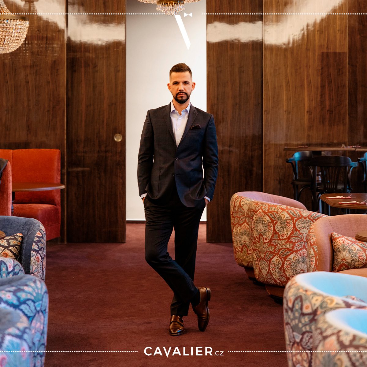 Muž stojící v elegantním hotelovém salonku v tmavě modrém dvoudílném obleku na míru Cavalier se světlou košilí bez kravaty – ukázka nadčasového formálního obleku vhodného pro večerní i business události.
