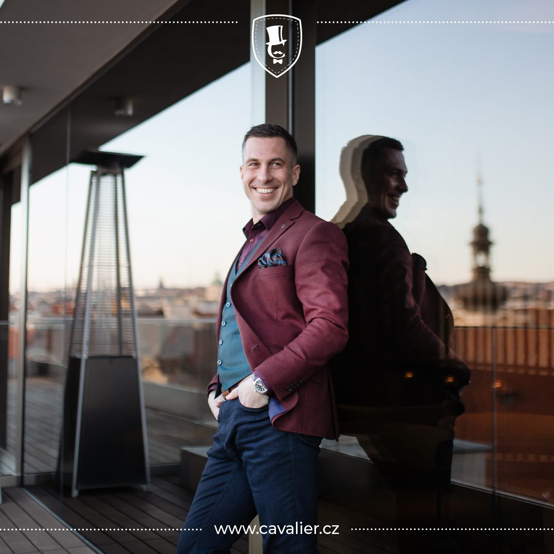 Usměvavý muž v moderním smart casual outfitu – vínové sako na míru Cavalier doplněné tmavě zelenou vestou, tmavou košilí a džíny představuje stylovou kombinaci výrazných barev a strukturovaných materiálů.