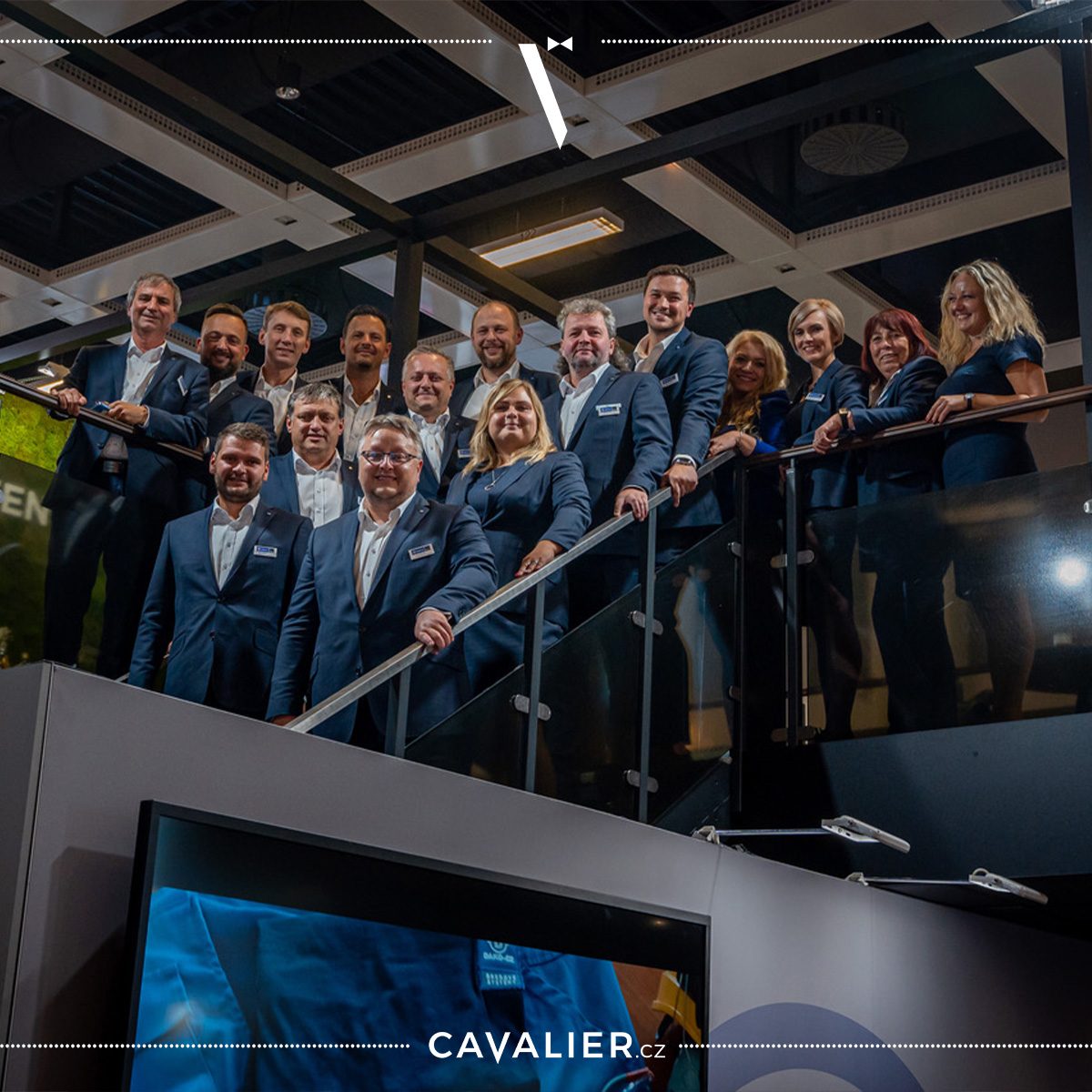 Skupinová fotografie týmu v elegantních tmavě modrých kostýmech a oblecích na míru od Cavalier na schodišti – ukázka reprezentativního firemního oblečení pro české týmy.