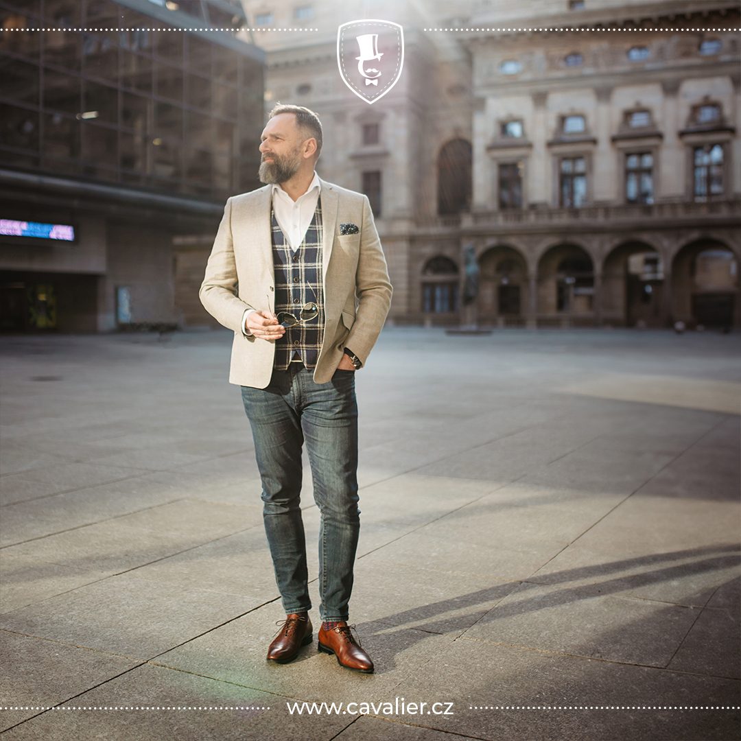 Muž v dokonalém smart casual outfitu – světlé vlněné sako na míru Cavalier zkombinované s kostkovanou vestou, bílou košilí bez kravaty a tmavými džíny.