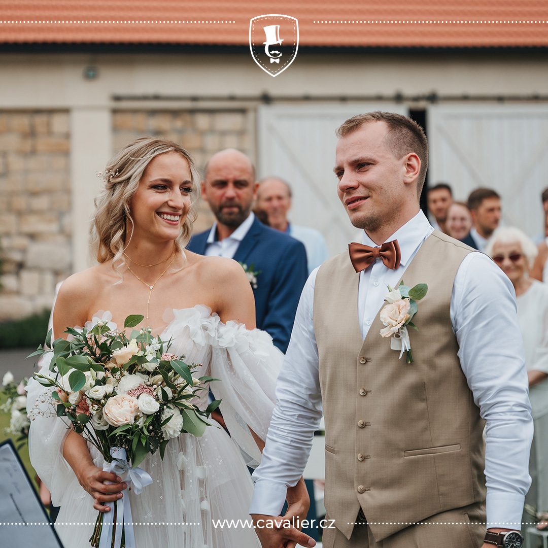 ženich v béžové vestě na míru Cavalier s hnědým motýlkem a nevěsta s kyticí jako ukázka dokonale sladěného outfitu na svatbu ve stodole