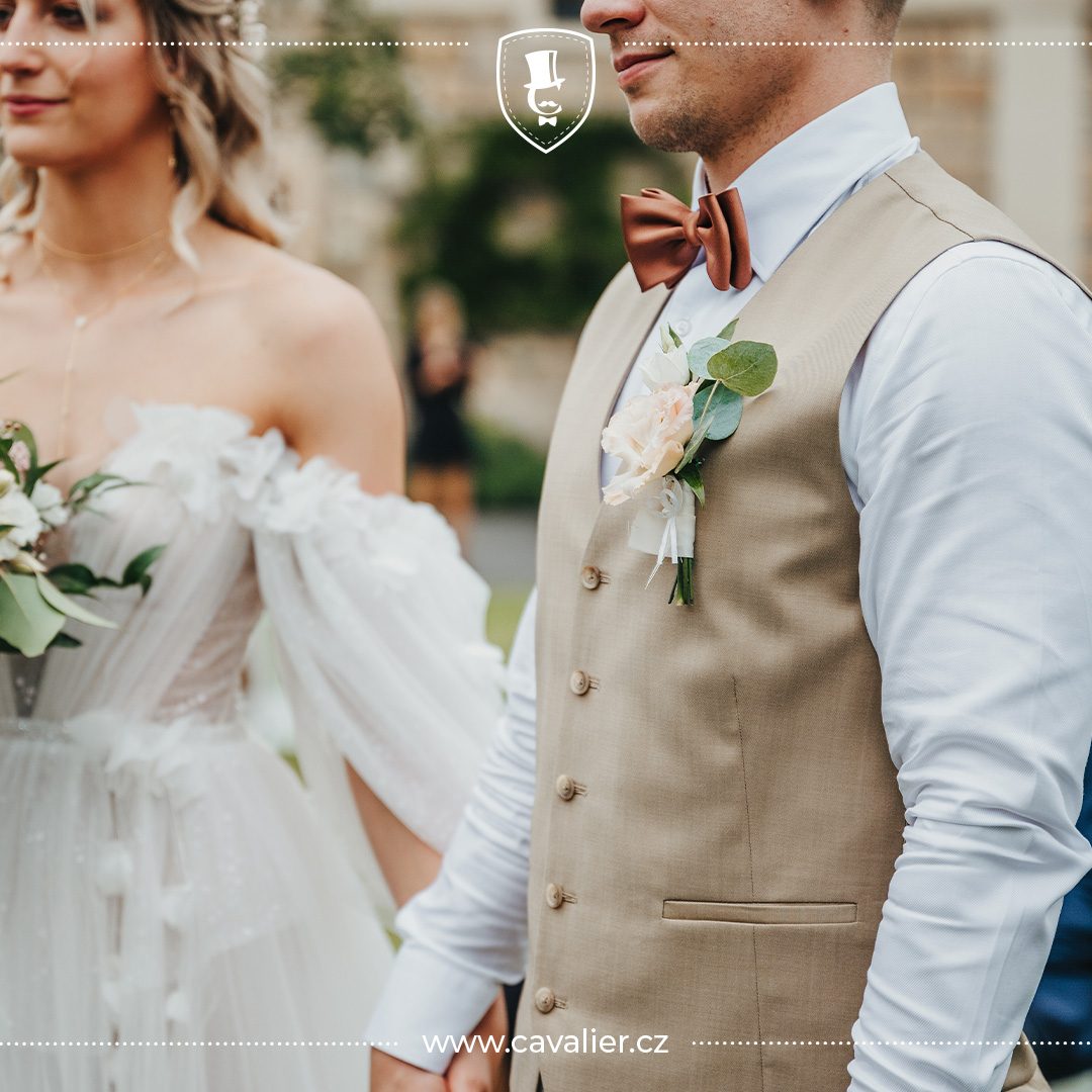 Detail ženicha v béžové vestě na míru Cavalier s hnědým motýlkem a kytičkou v klopě pro dokonale sladěný outfit na svatbu ve stodole