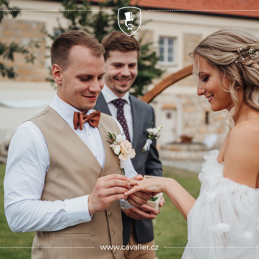 Ženich v pískové vestě na míru Cavalier nasazuje prsten nevěstě, zatímco svědek v elegantním tmavě šedém obleku s kravatou přihlíží v pozadí