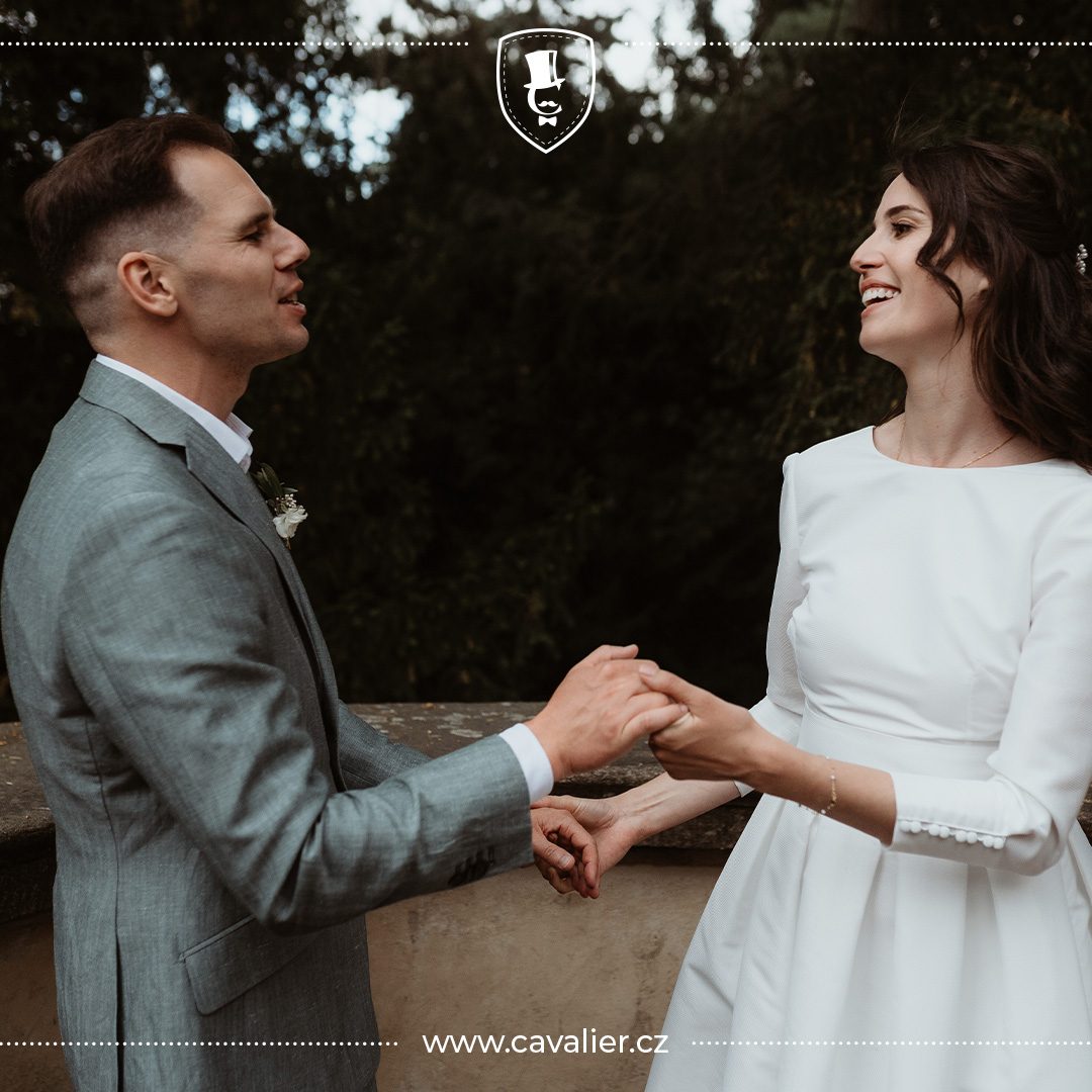 Romantický pohled novomanželů držíce se za ruce a ženich v šedozeleném saku na míru Cavalier jako ukázka dokonale sladěného svatebního stylu